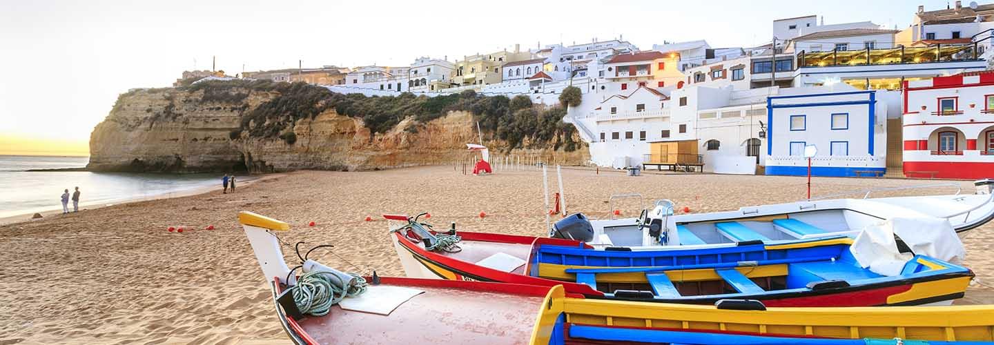 In the foreground the colourful boats on a beach, in the background the white houses with the colourful boride of Faro in Portugal.