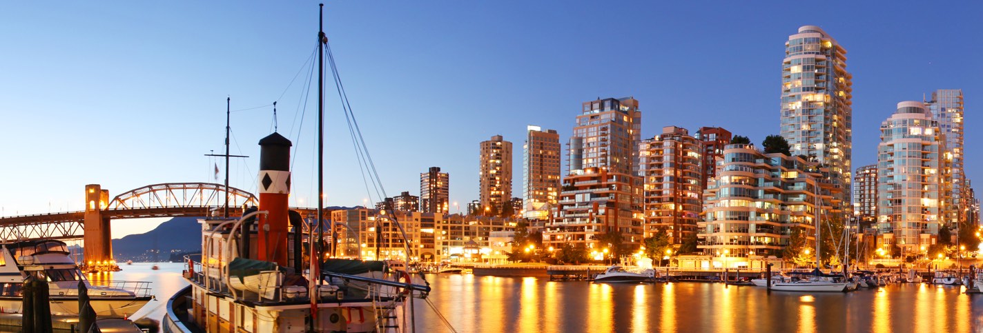 Photo of the city of vancouver at night with its illuminated skyscrapers in the back, and a river with boats in the front.