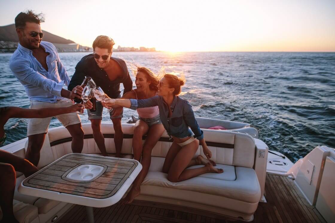 Group of young people sitting at the back of a yacht and toasting drinks. Young people having a boat party at sunset.
