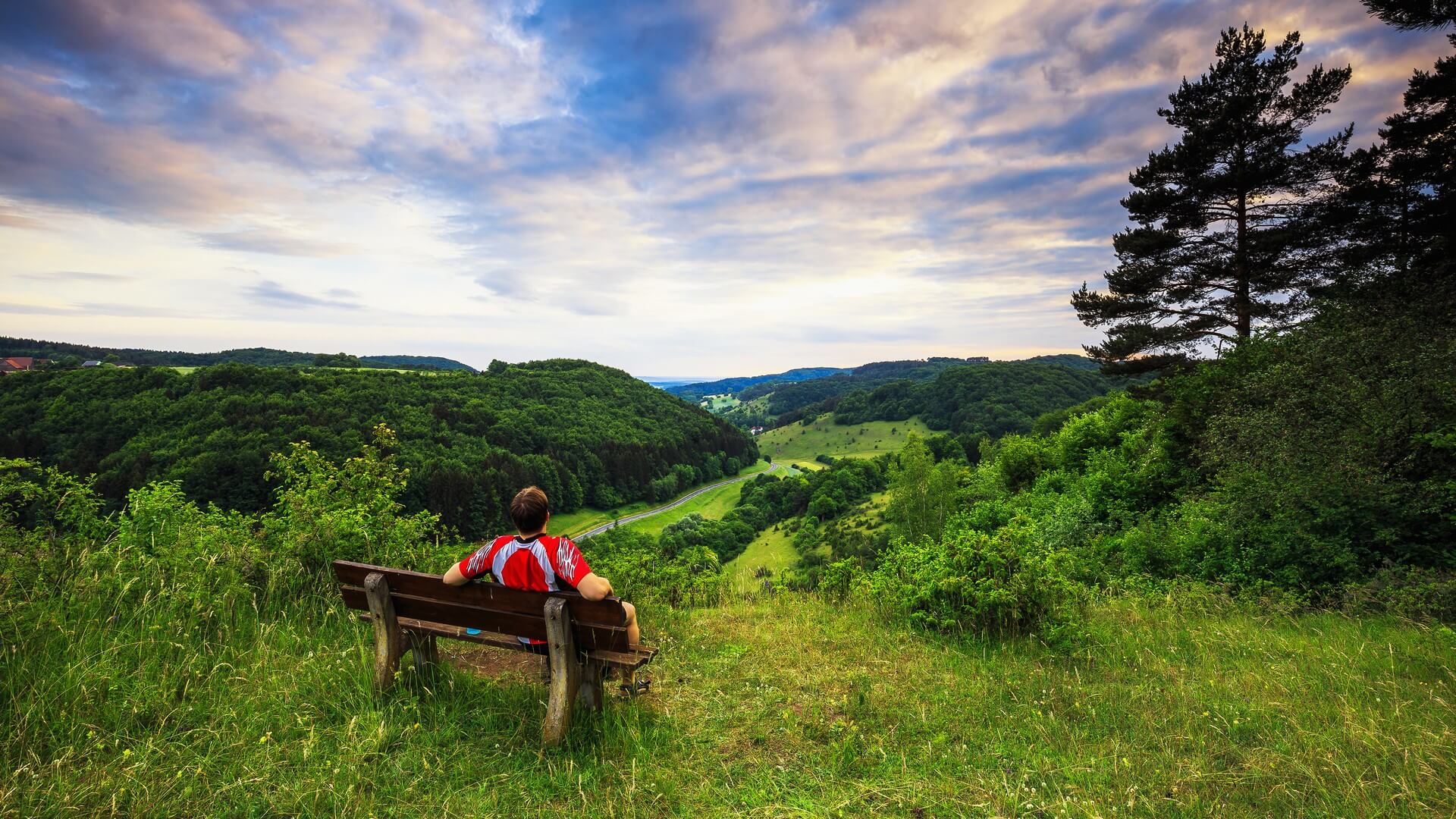 Junger Mann im roten Hemd auf einer Bank, den atemberaubenden Blick auf eine frühsommerliche bayerische Hügellandschaft in Franken, Deutschland genießend
