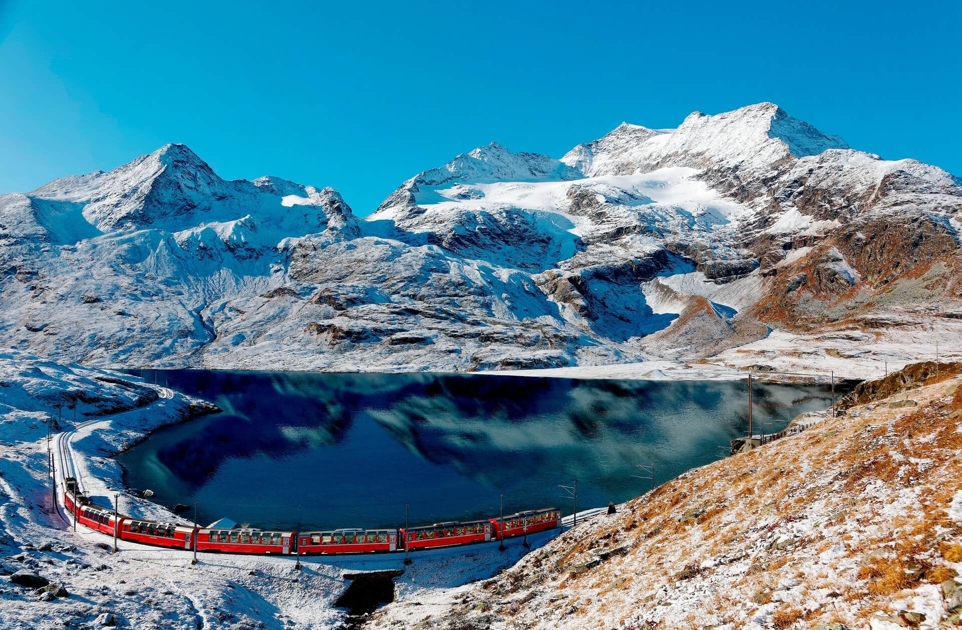 A Bernina Express train traveling along the lake shore of Lago Bianco & alpine mountains towering under blue sky in background after a snowfall in autumn, near Ospizio Bernina, in Grisons, Switzerland
