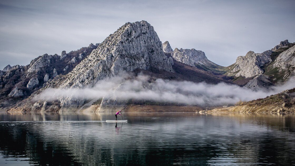 A girl or woman with pink coat practices paddle surfing exploring in a mountain lake with foggy
