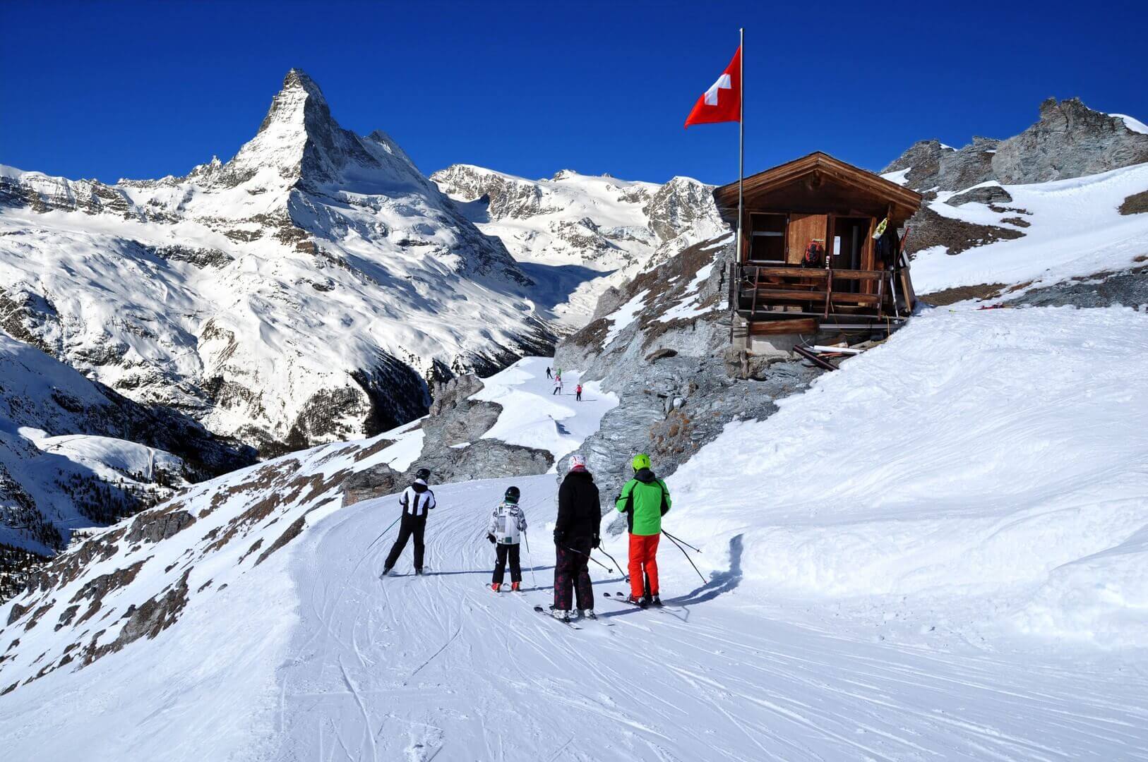 Skieurs sur le chemin de la piste de ski dans les Alpes suisses en journée ensoleillée, derrière le Cervin. Petite maison en bois avec drapeau suisse rouge à proximité de la piste de ski. Manipulation de photos de montagnes et de stations de ski, Suisse.