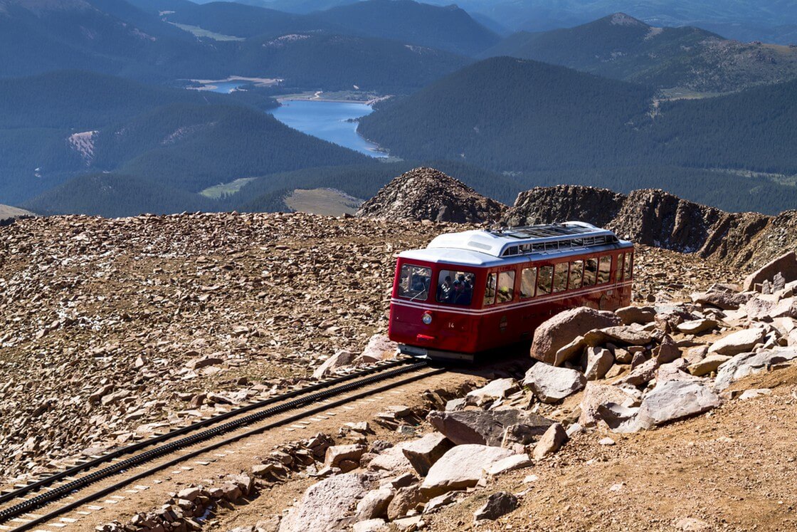 Pikes Peak Cog Railway car traveling down from top of Pikes Peak mountain top in Colorado on sunny summer morning with mountains and lake in distance
