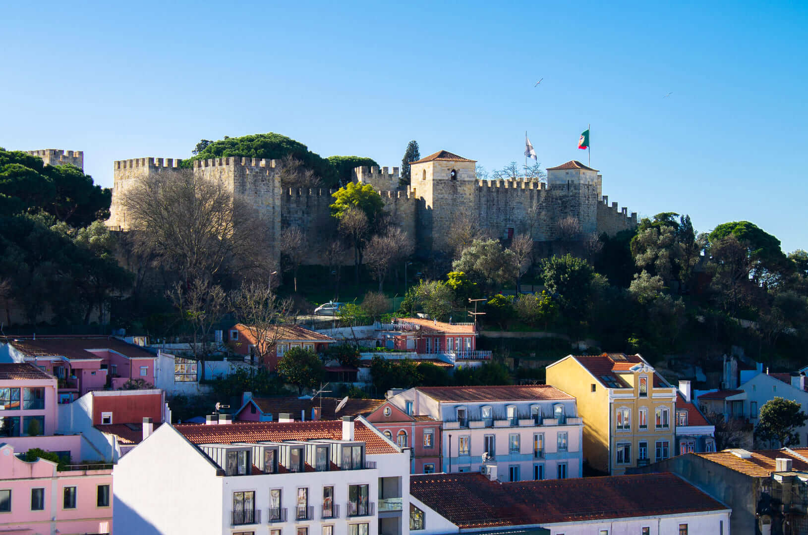 A view across the colourful buildings, rooftops and gardens of Lisbon, Portugal, towards the imposing Castelo de S. Jorge (St George Castle) on a sunny day.
