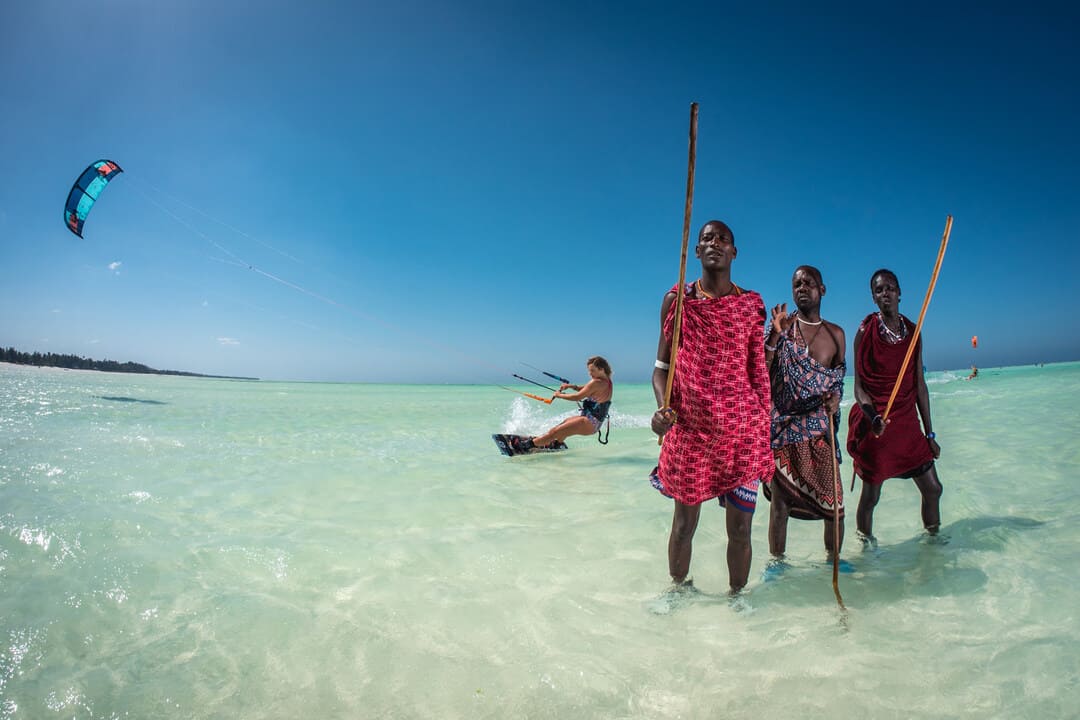 Eine Frau kitesurft am Strand von Sansibar, während 3 Inder vom Wasser aus in die Kamera schauen.