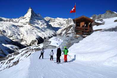 Skiers on the way to ski slope in Swiss Alps in sunny day, Matterhorn behind. Small wooden house with red swiss flag near by ski slope. Mountains and skiing resort photo manipulation, Switzerland.
