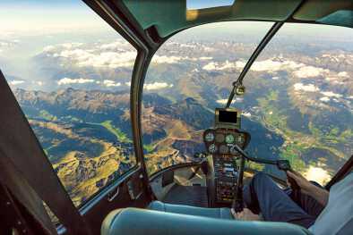 Cabina de helicóptero volando sobre paisajes montañosos y cielo nublado, con brazo piloto conduciendo en cabina. Espectacular vista aérea de los Alpes.