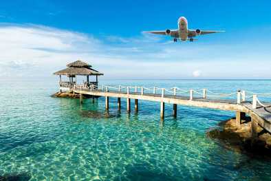 Un jet privé volant près d'un pont menant vers un petit kiosque, au-dessus de l’eau turquoise, sur une île exotique