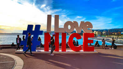 Niza, Francia: Turistas tomando fotos en el cartel "I love Nice" con vista del paisaje urbano de Niza, el Mediterráneo y el Paseo de los Ingleses.