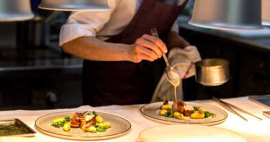 Chef preparing a plate made of meat and vegetables. The chef is pouring sauce on two plates.