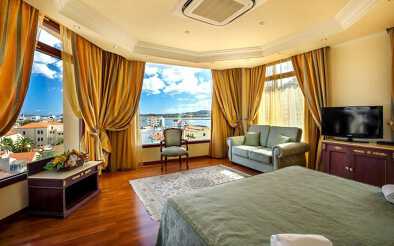 Photographie d'une chambre colorée et luxueuse de l'hôtel Panorama à Olbia en Sardaigne. Les rideaux de couleur orange sont ouverts autour des 3 grandes fenêtres laissent entrevoir la superbe vue panoramique sur la ville.
