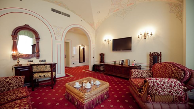 The living room of this opulent suite in appointed in distinctive Duke’s palace style furnished with massive but extremely comfortable armchairs, tassel-fringed chaise longue with magnificent crimson, pelmet-topped drapes frame large floor to ceiling windows leading to a balcony overlooking the street below.

