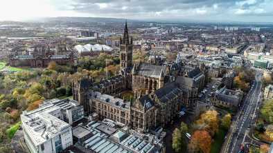 Imagen aérea de bajo nivel sobre el follaje otoñal de los árboles en Kelvingrove Park, Glasgow, hasta la torre gótica de la Universidad de Glasgow con el paisaje urbano detrás.