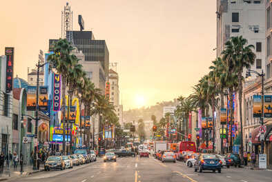 Einkaufsstraße in Los Angeles auf dem Sunset Strip, gesäumt von Geschäftsschildern, Palmen, Verkehr und dem Sonnenuntergang im Hintergrund.