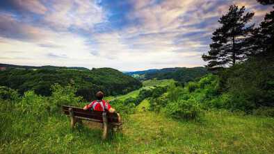 Joven con camisa roja en un banco, disfrutando de las impresionantes vistas de un paisaje de colinas bávaras de principios de verano en Franconia, Alemania