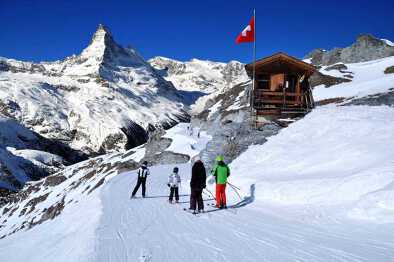 Sciatori sulla strada per la pista da sci nelle Alpi svizzere in una giornata di sole, con il Cervino alle spalle. Piccola casa di legno con bandiera svizzera rossa vicino alla pista da sci. Manipolazione fotografica di montagne e stazioni sciistiche, Svizzera.
